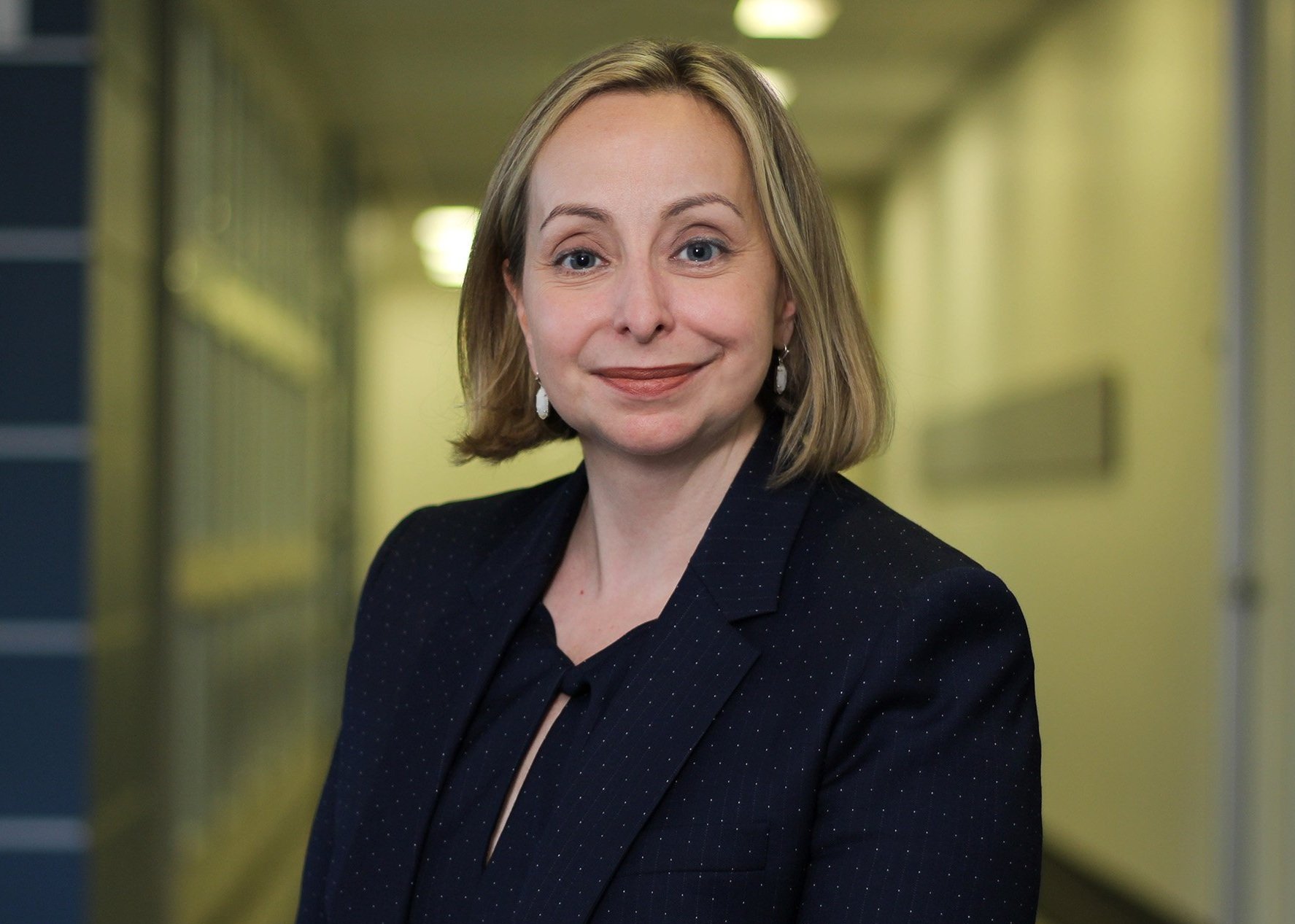 A woman with shoulder-length blonde hair, wearing a black blazer with subtle dots, standing in a brightly lit corridor with yellow and blue walls.