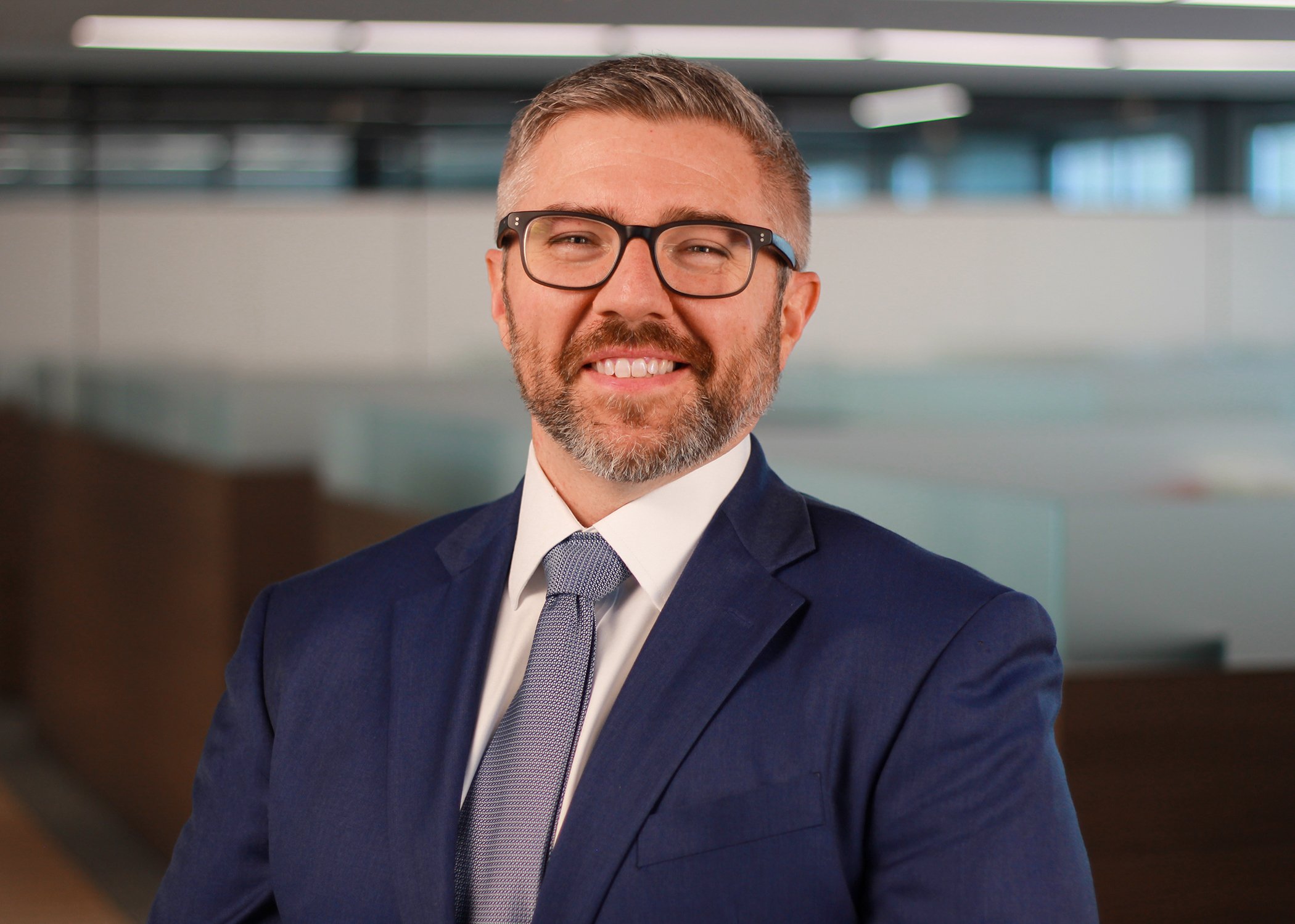 A middle-aged man with short gray hair, glasses, and a beard, wearing a navy blue suit, white shirt, and gray tie, smiling confidently in a modern office environment.