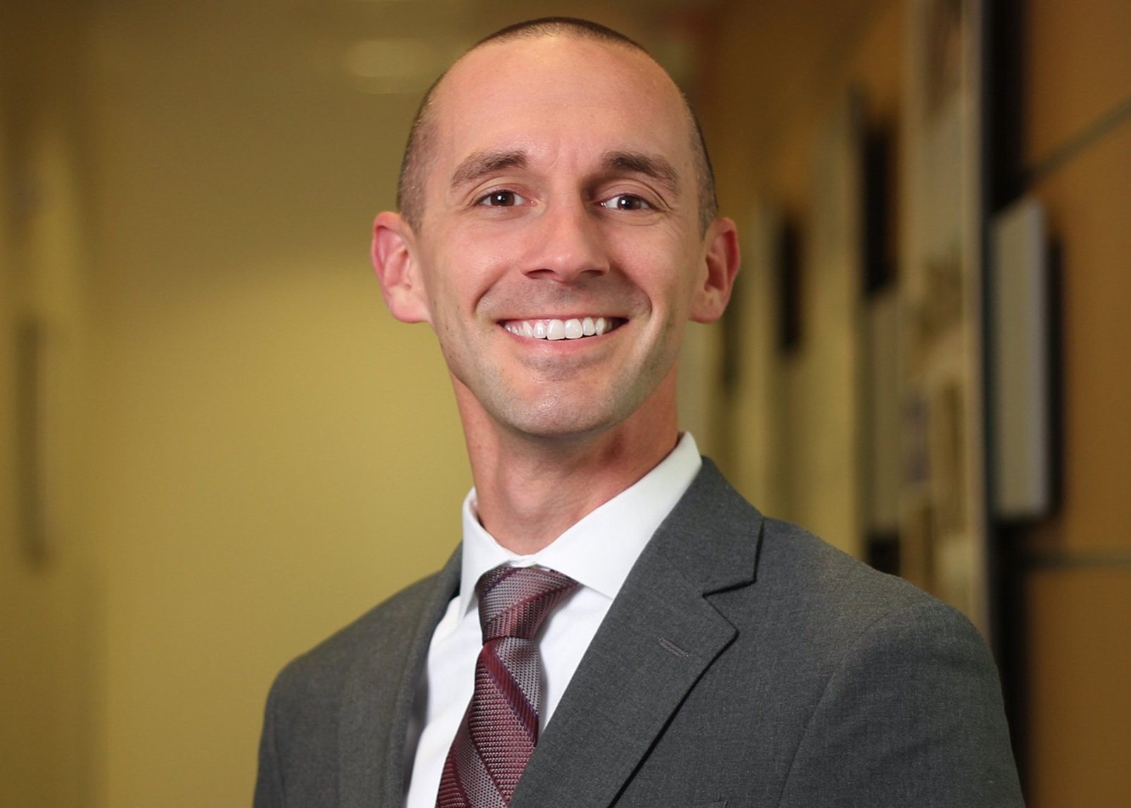 A man in a gray suit, white shirt, and maroon striped tie smiling in an indoor hallway with framed pictures or artwork on the wall.