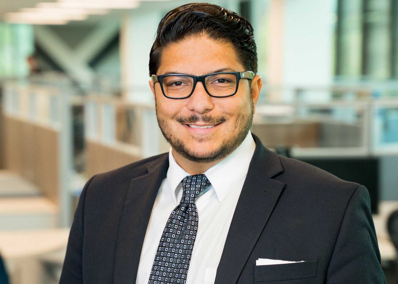 A professional man with dark hair, glasses, and a beard, wearing a black suit, white shirt, and patterned tie, smiling in an office setting.